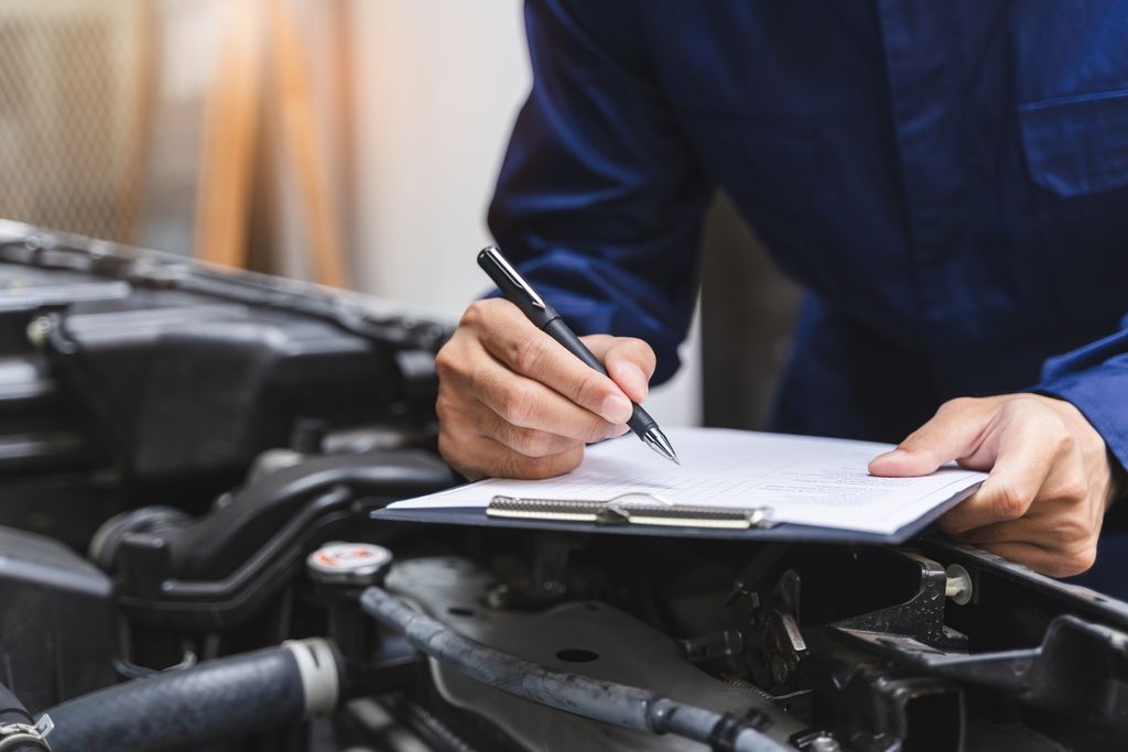 A mechanic writes on a clipboard while inspecting a car engine.