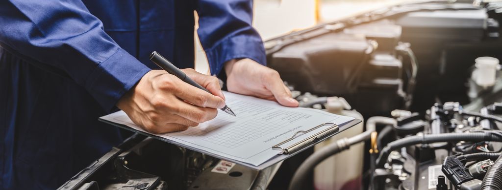 A mechanic in blue overalls writes on a clipboard while inspecting a car engine.